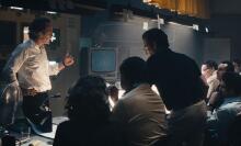 People sit and stand in a newsroom, talking and looking at one another.