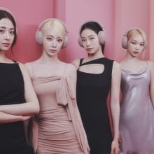 The members of Le Sserafim wearing Sony headphones and standing in front a pink wall.